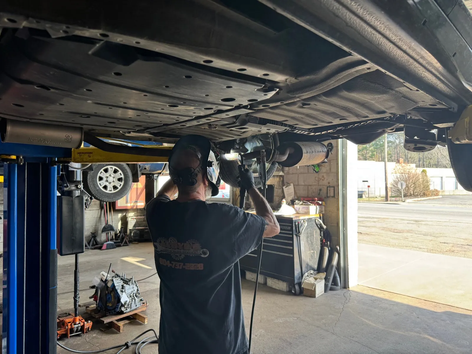 A technician working overhead on the exhaust of a vehicle raised on the lift, with the open bay door behind him