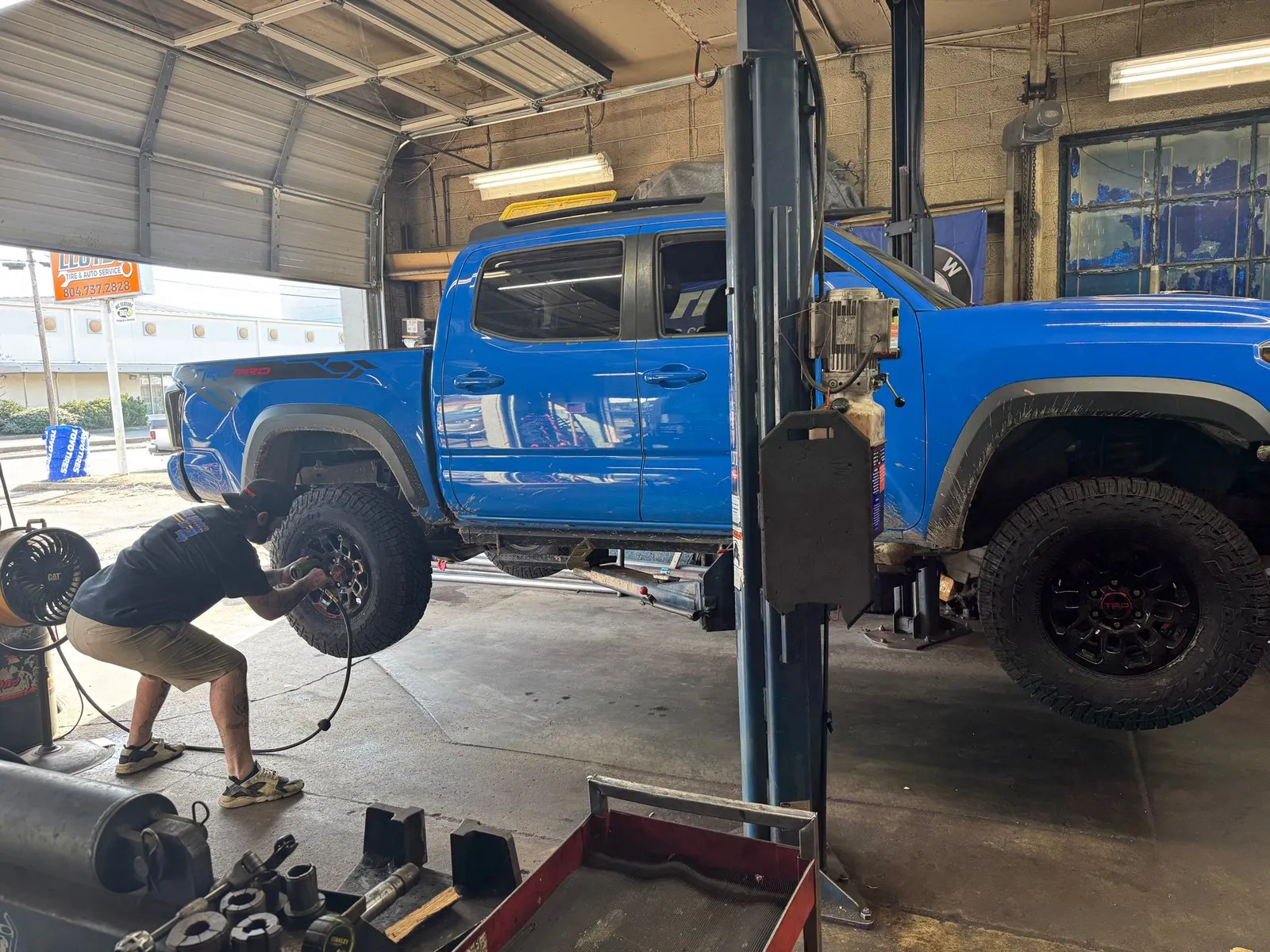 A blue Toyota Tacoma raised on the two-post lift while a technician runs an impact wrench on the front wheel