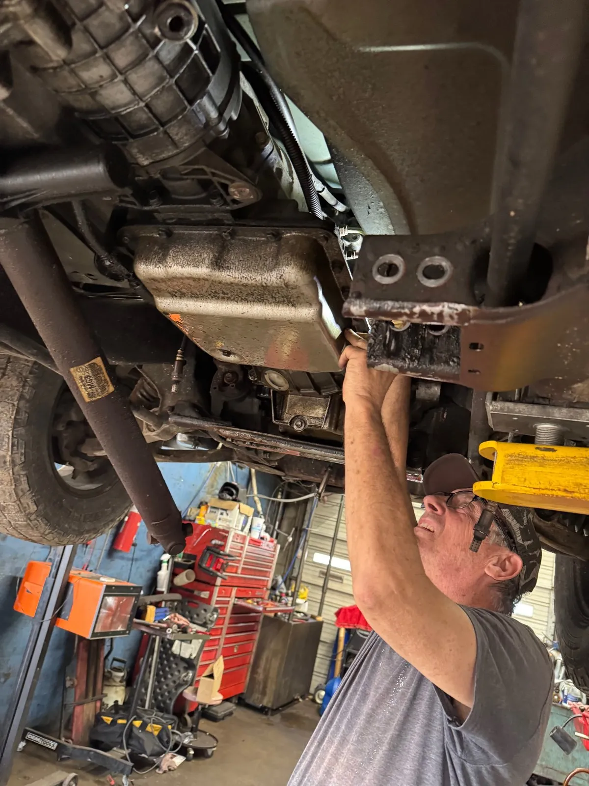 A technician reaching up under a raised vehicle to work on the transmission crossmember