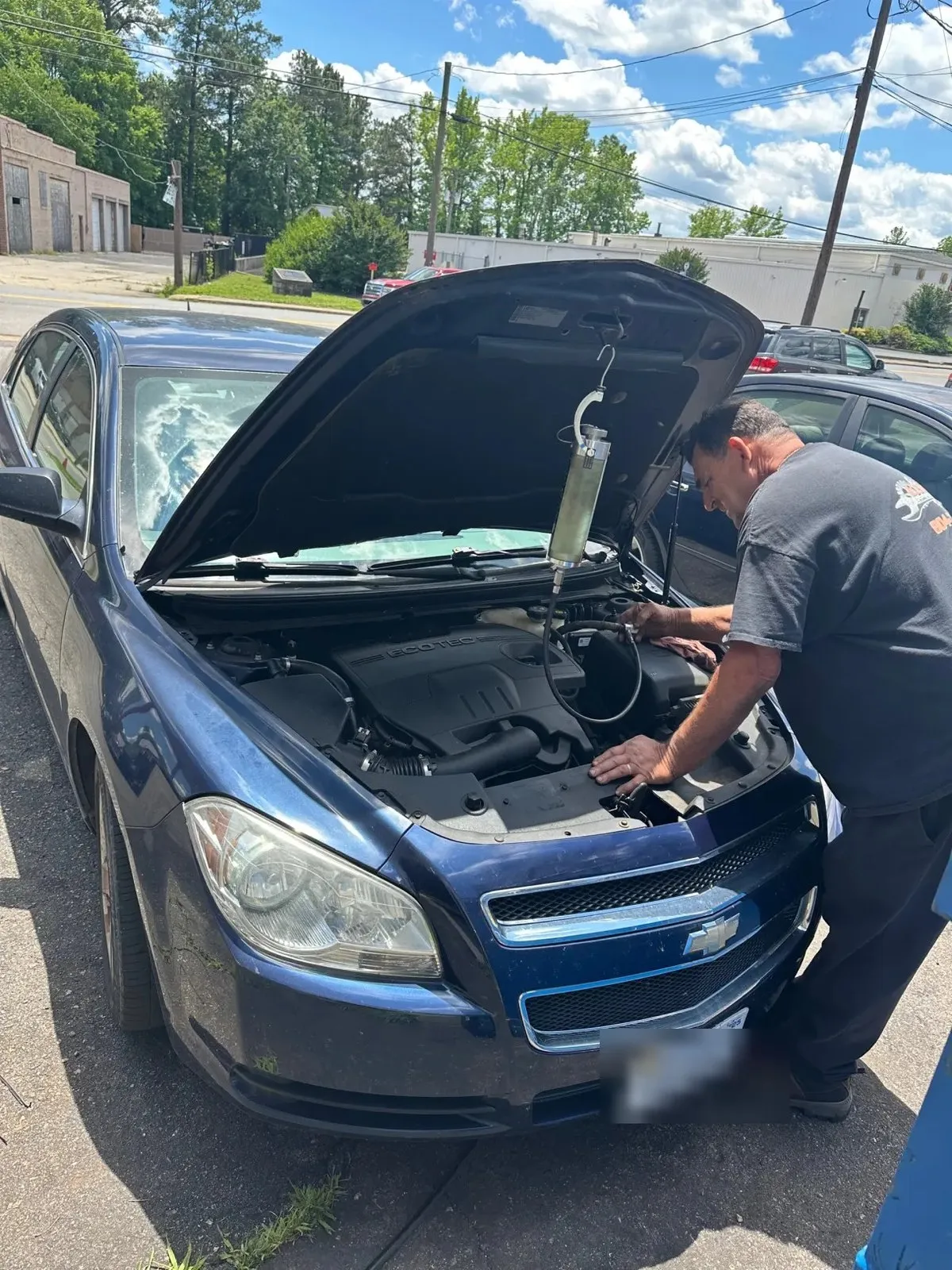 A technician working under the open hood of a blue Chevrolet Malibu on the lot, with a cooling-system tester fitted